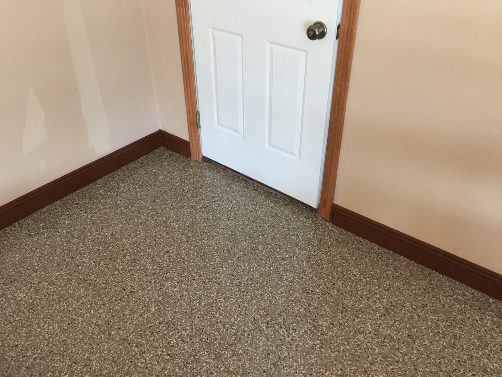 An interior corner of a room with light peach walls, brown wooden baseboards, and a speckled grey floor.