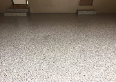 A garage floor with a grey and white speckled epoxy finish, leading to two small sets of stairs against a beige wall.