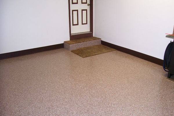 A room with a speckled epoxy floor, white walls, dark trim, and a small step leading to a white door.