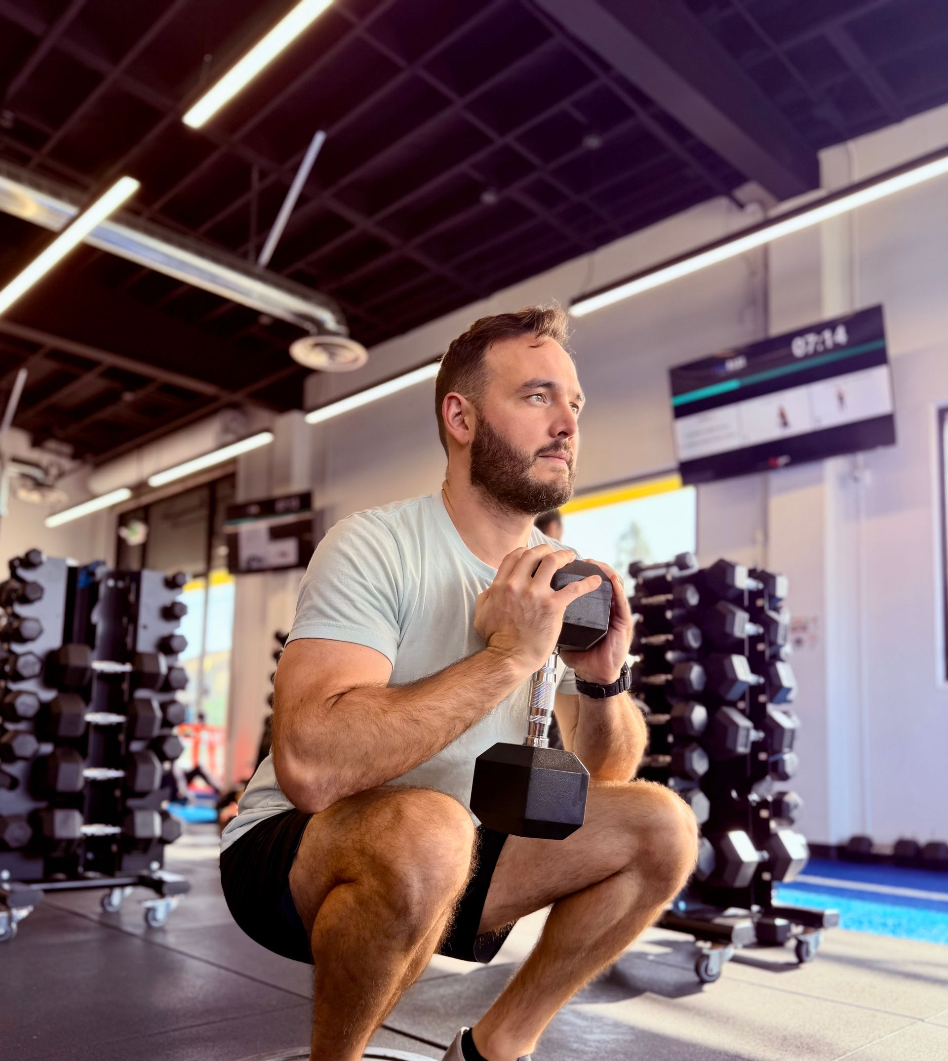 Man in gym holding dumbbell, squatting low. Black shorts, light green shirt.