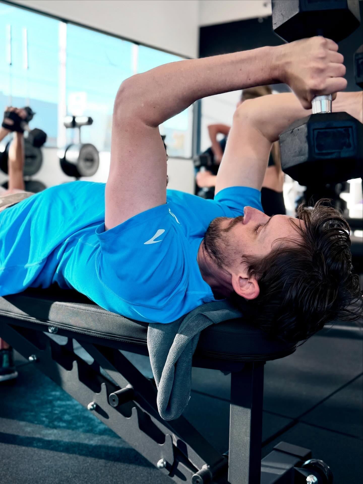 Man on a bench presses a dumbbell in a gym. He wears a blue shirt.