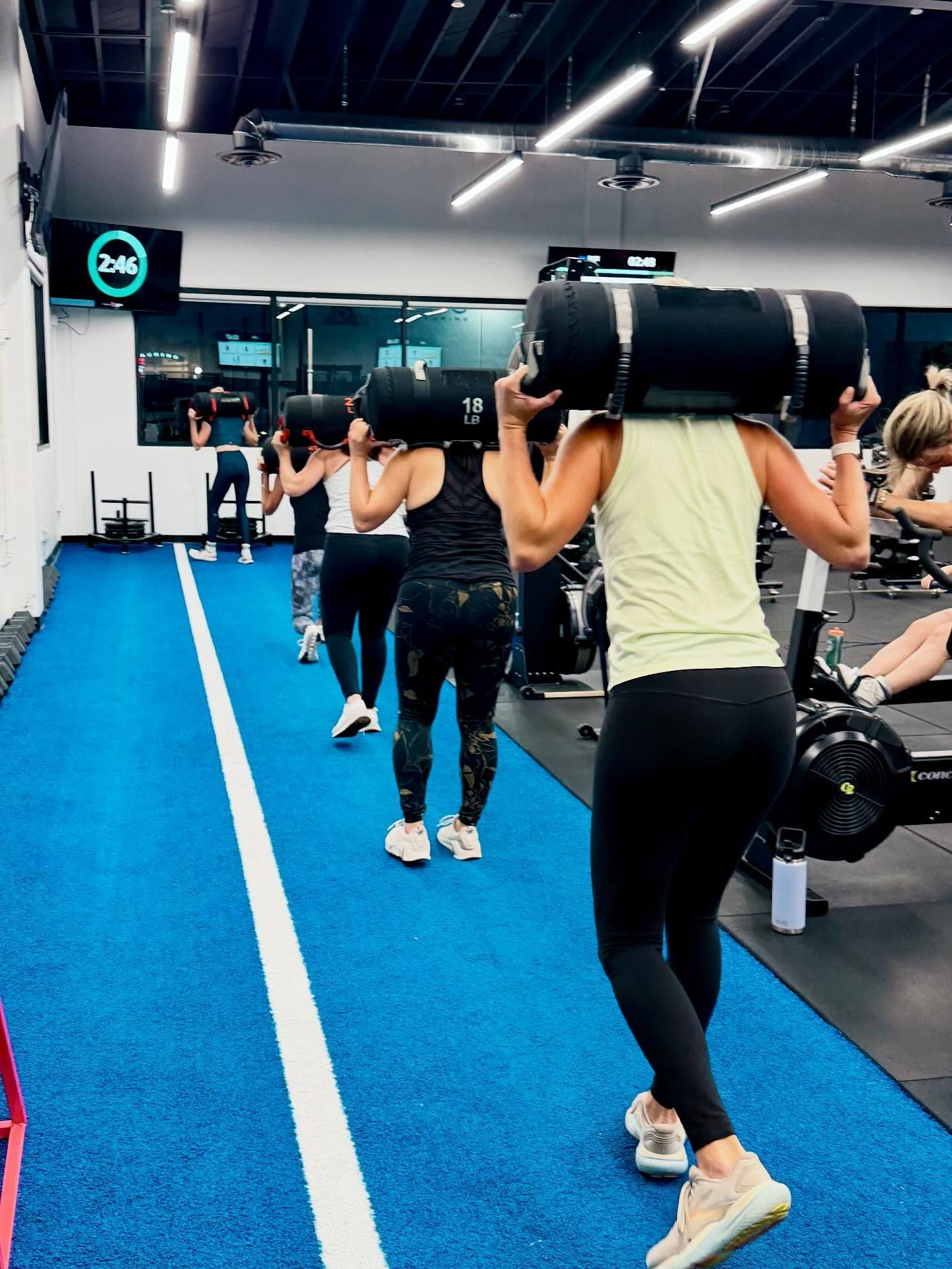 People exercising with weights at a gym; blue floor, black weights, person in a lime green shirt.