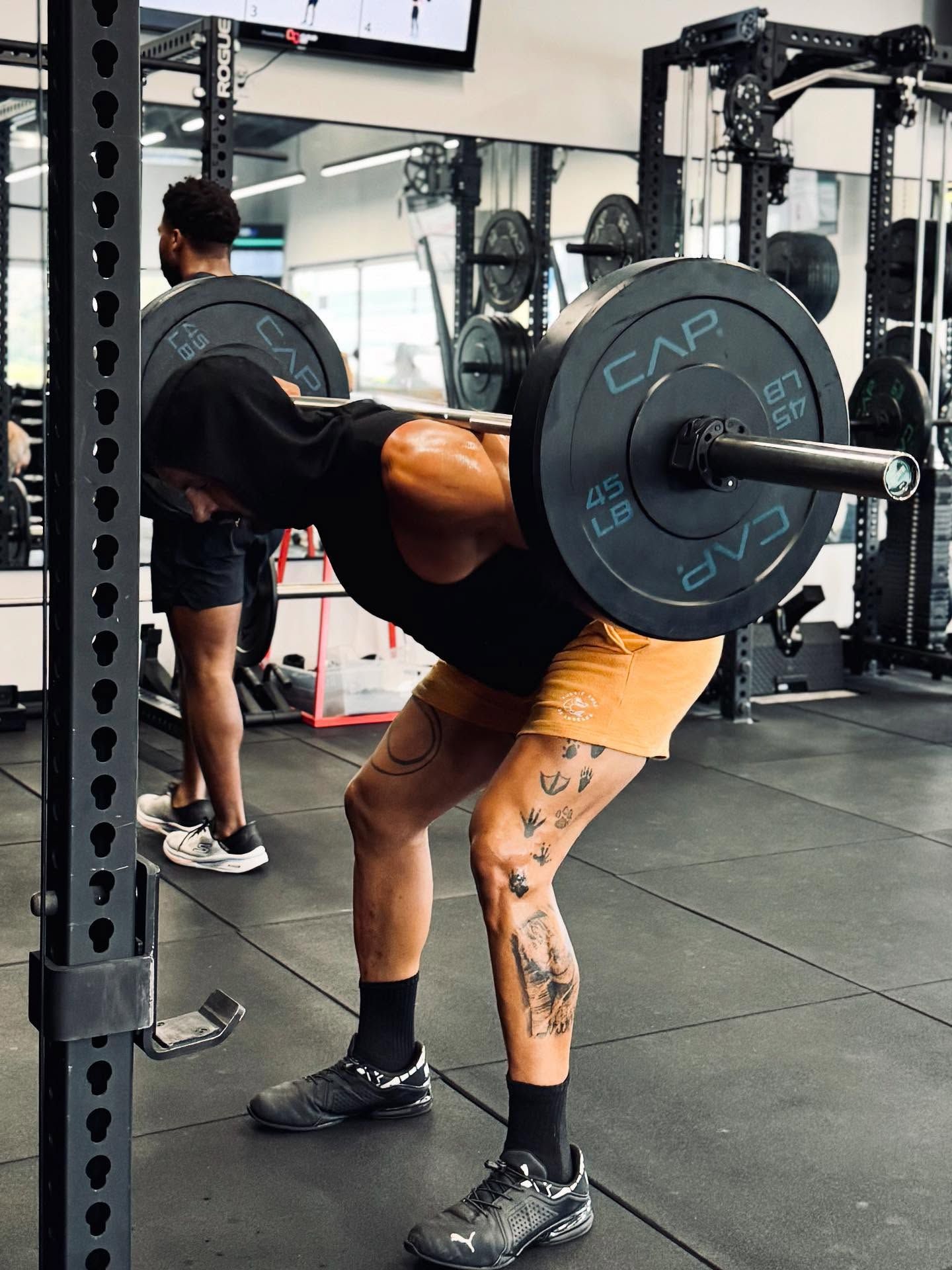 Person performing a barbell squat in a gym. Black weight plates on the bar. Tattoos on the person's leg.