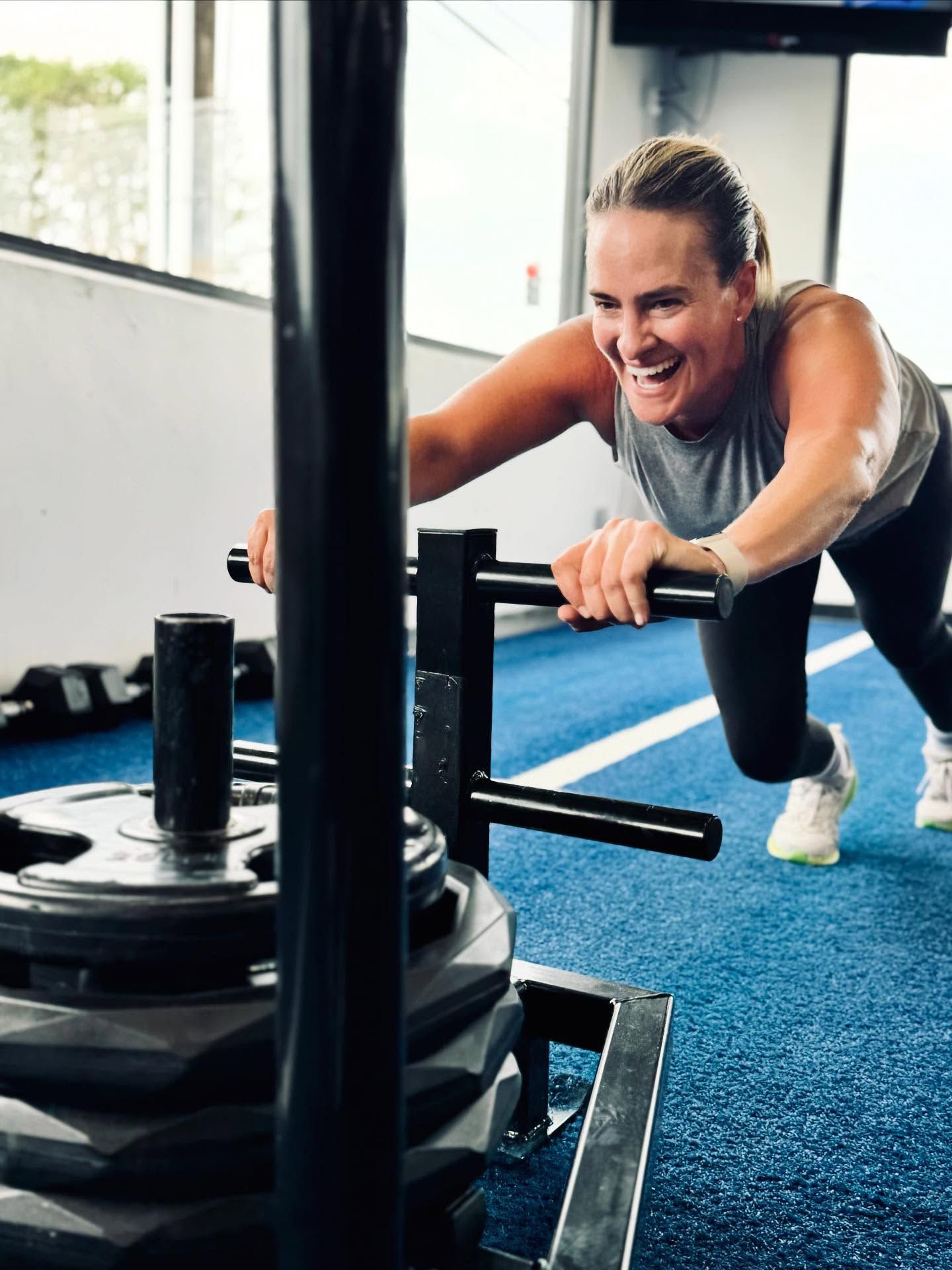 Woman pushing a weighted sled at a gym, smiling, wearing athletic wear on a blue mat.