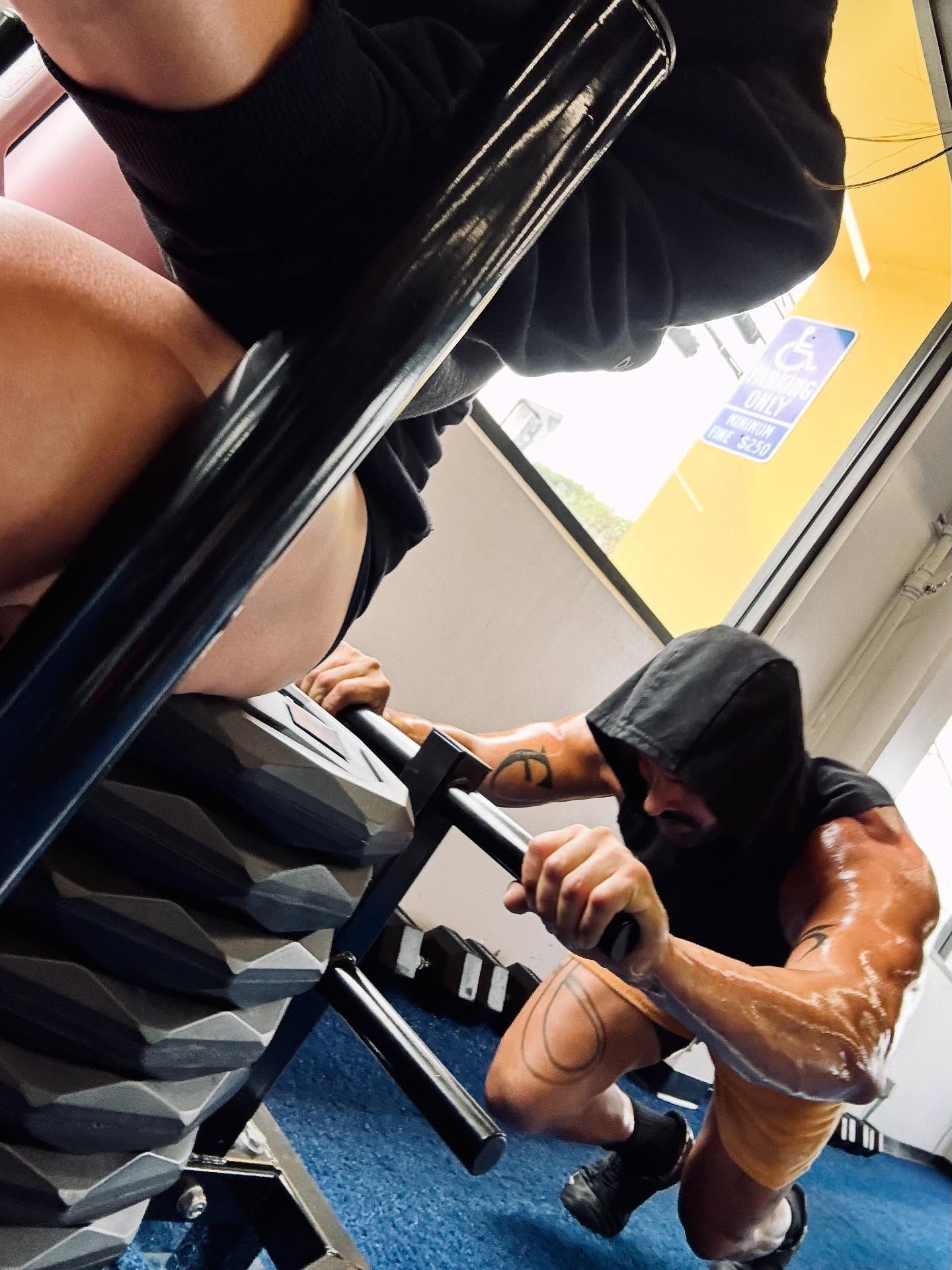 Man exercising on a stair-climbing machine. He is wearing a black hoodie and has arm tattoos. Gym setting.