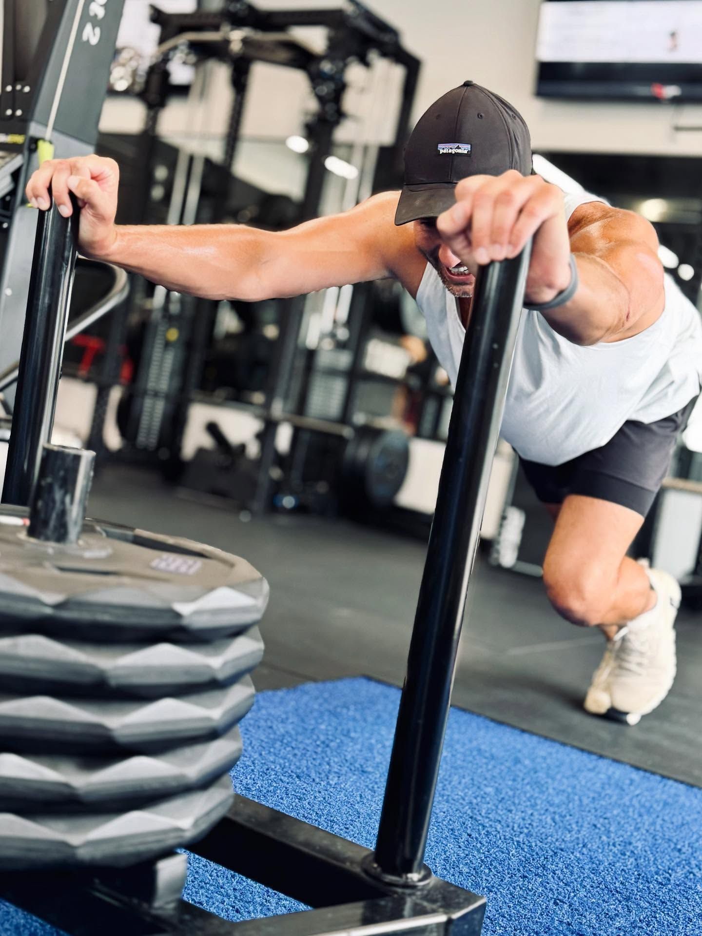 Man in a gym pushing a weighted sled, wearing a cap and athletic clothes.