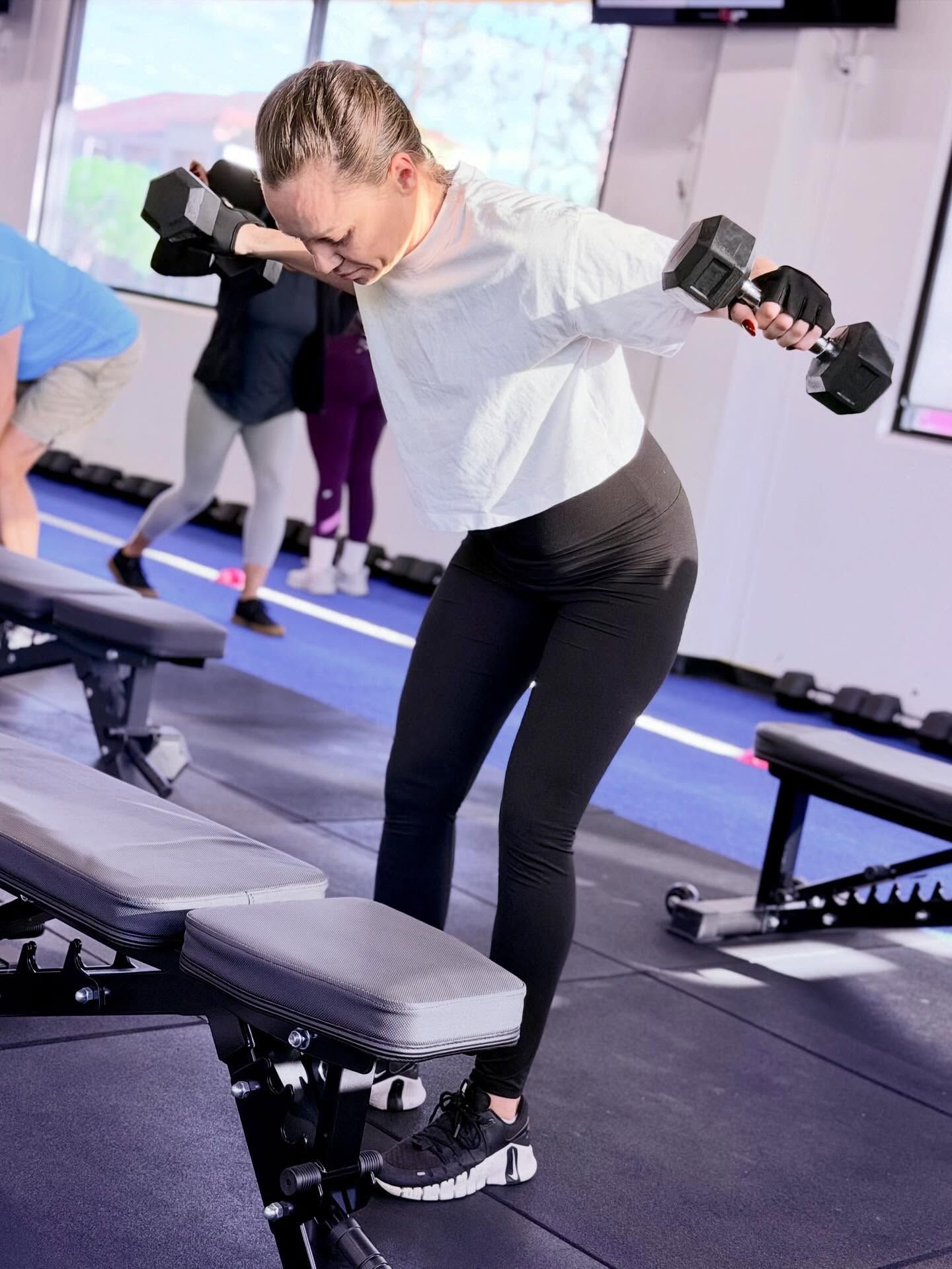 Woman exercising with dumbbells, leaning over a bench. Gym setting.