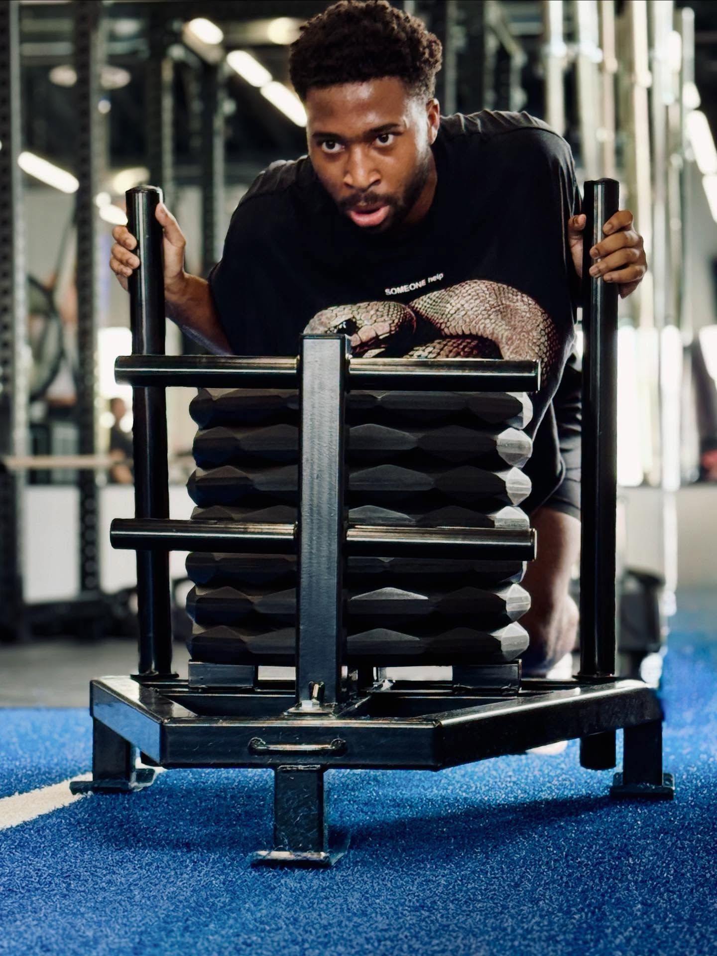 Man pushing a weight sled in a gym. Black shirt, intense expression, blue turf.