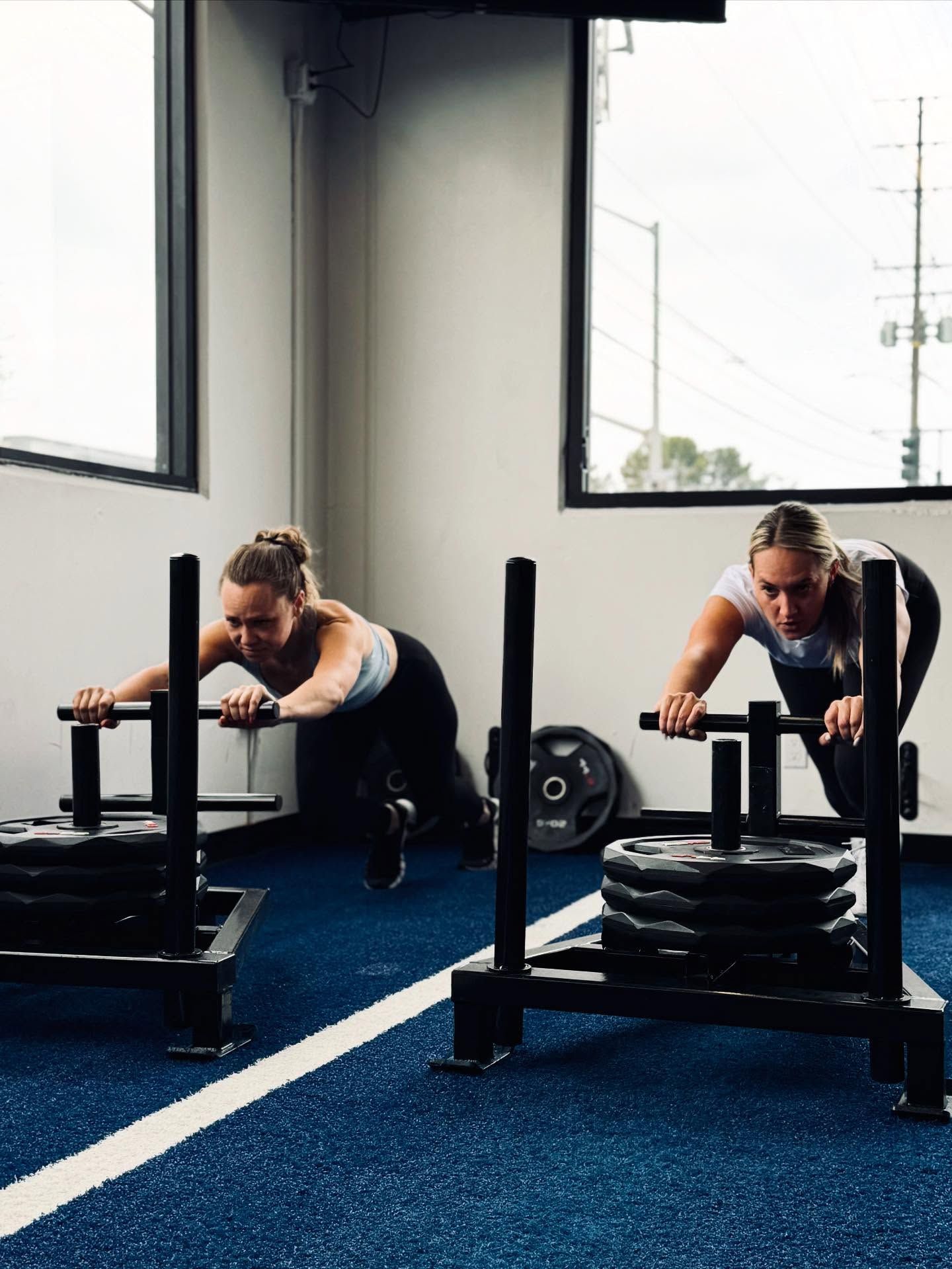 Two women push weighted sleds on a blue gym floor, near a window.