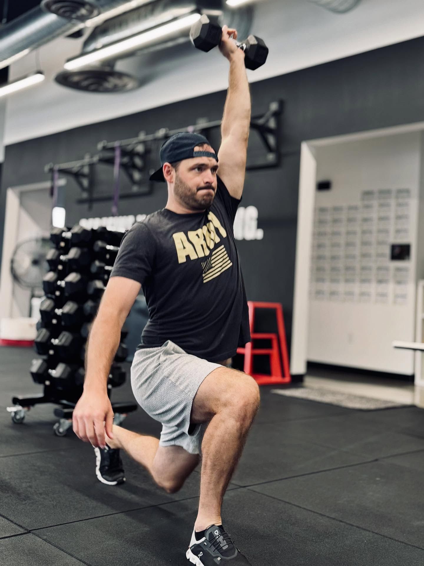 Man in gym doing a dumbbell overhead lunge, wearing a hat, shorts, and a t-shirt.
