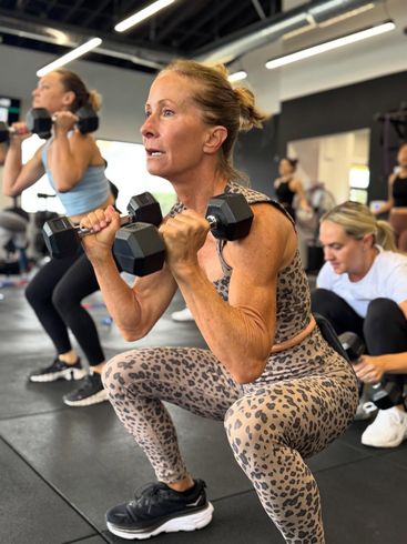 Woman in leopard print outfit doing dumbbell squats in a gym. Other people exercise in the background.