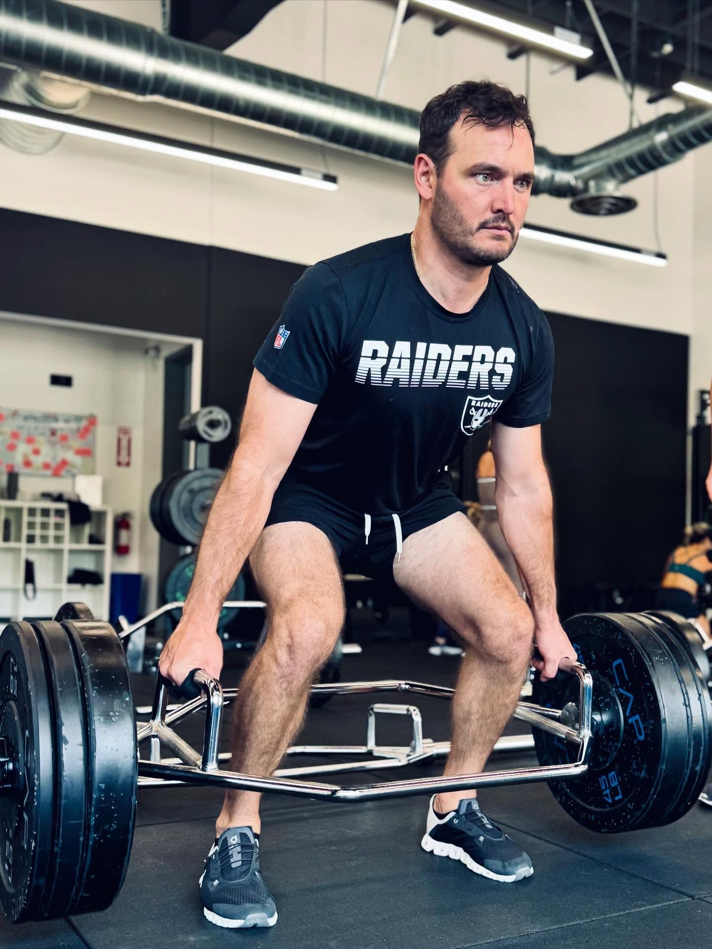 Man in Raiders shirt deadlifts a barbell in a gym.