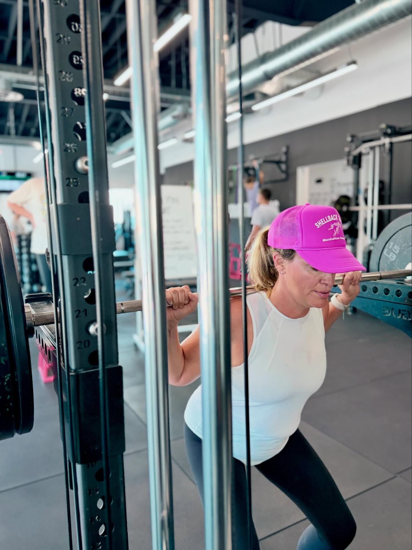 Woman in a white tank top, wearing a pink hat, squats with a barbell in a gym.