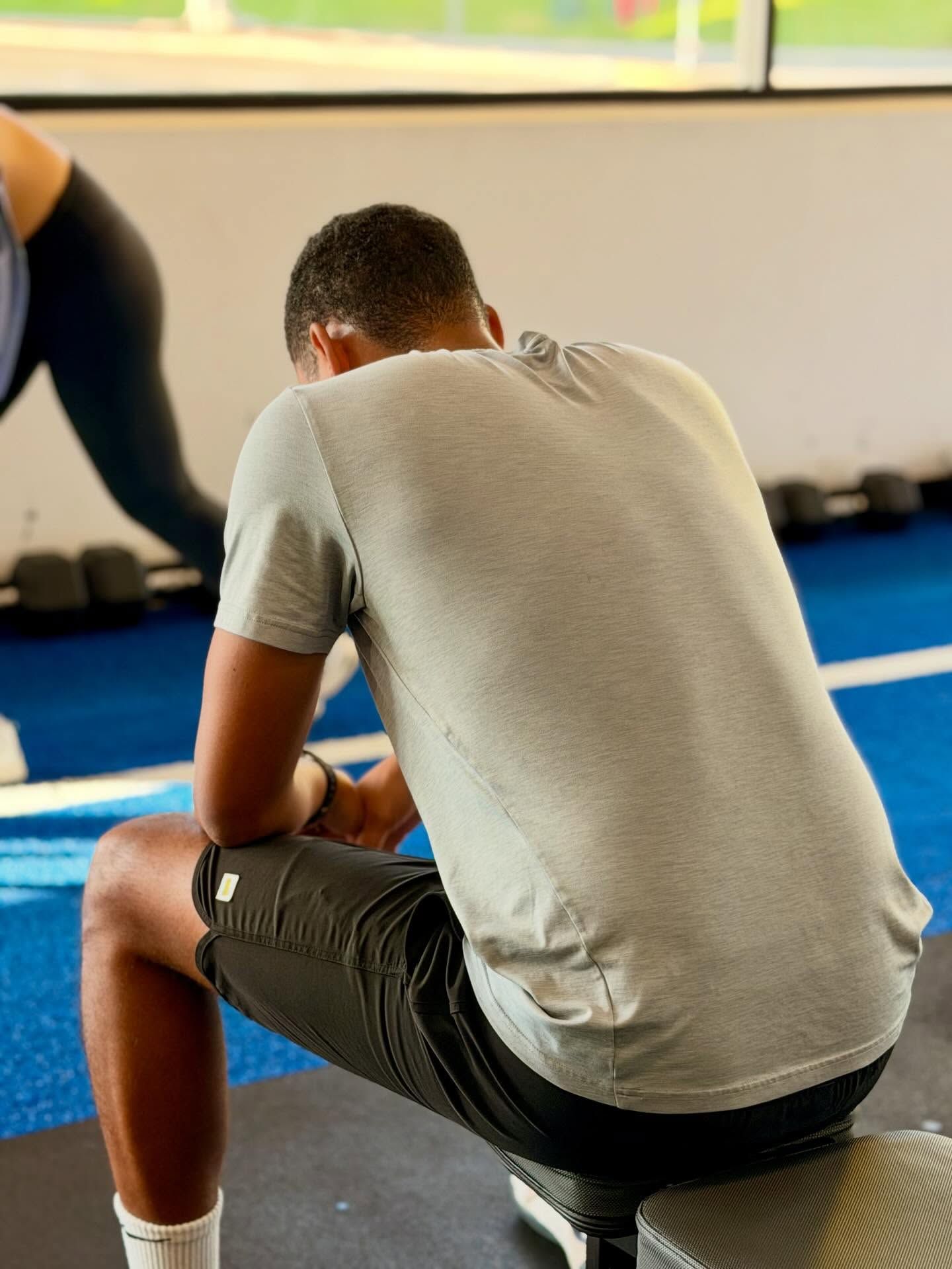 Man in gray shirt and black shorts sitting on a bench, looking down in a gym setting.