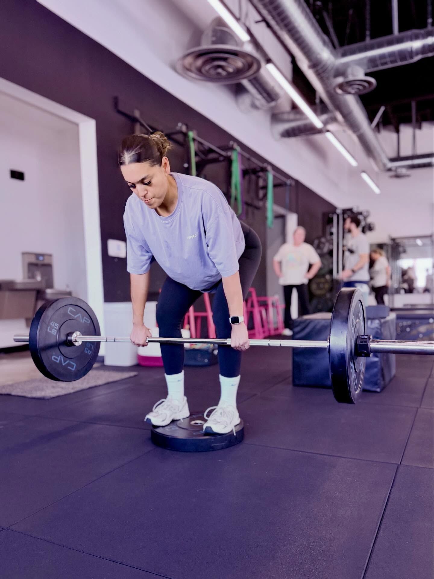 Woman in gym, lifting barbell off the floor on a platform. Purple floor, other people visible.