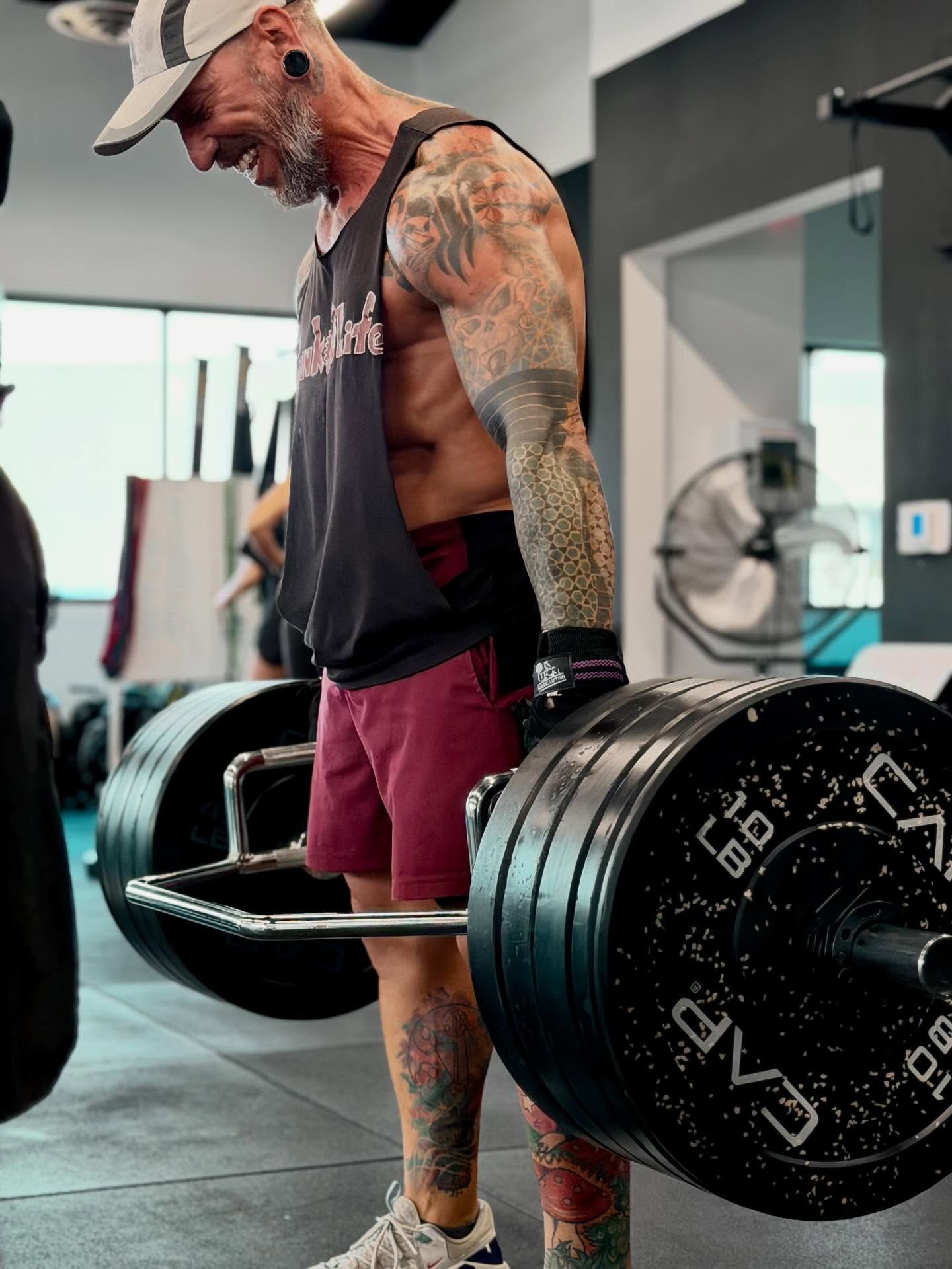 Man lifting heavy barbell in gym, wearing hat, tank top, shorts, and tattoos.