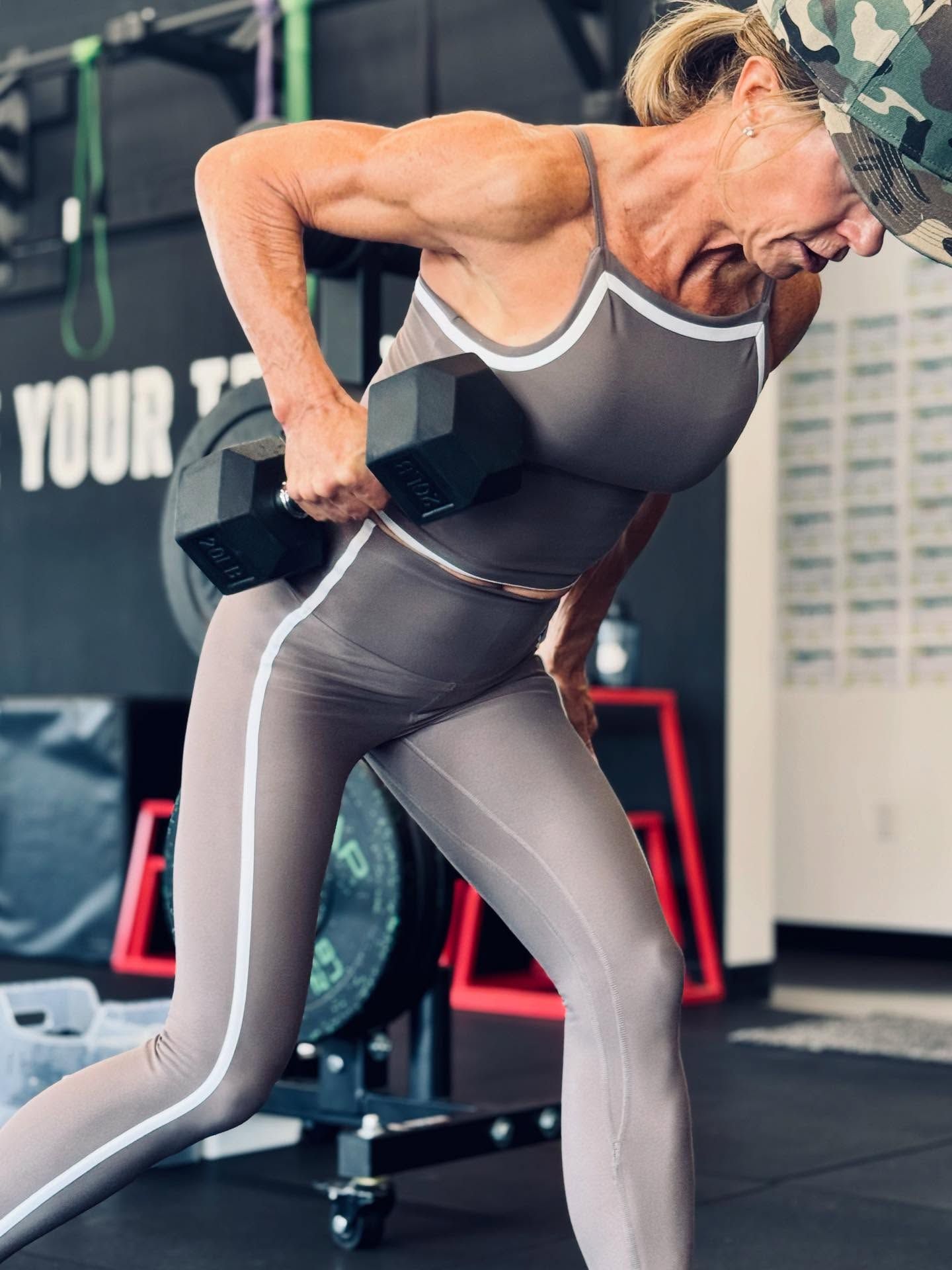 Woman in workout gear lifting a dumbbell, bent over in a gym, arm muscles flexed.