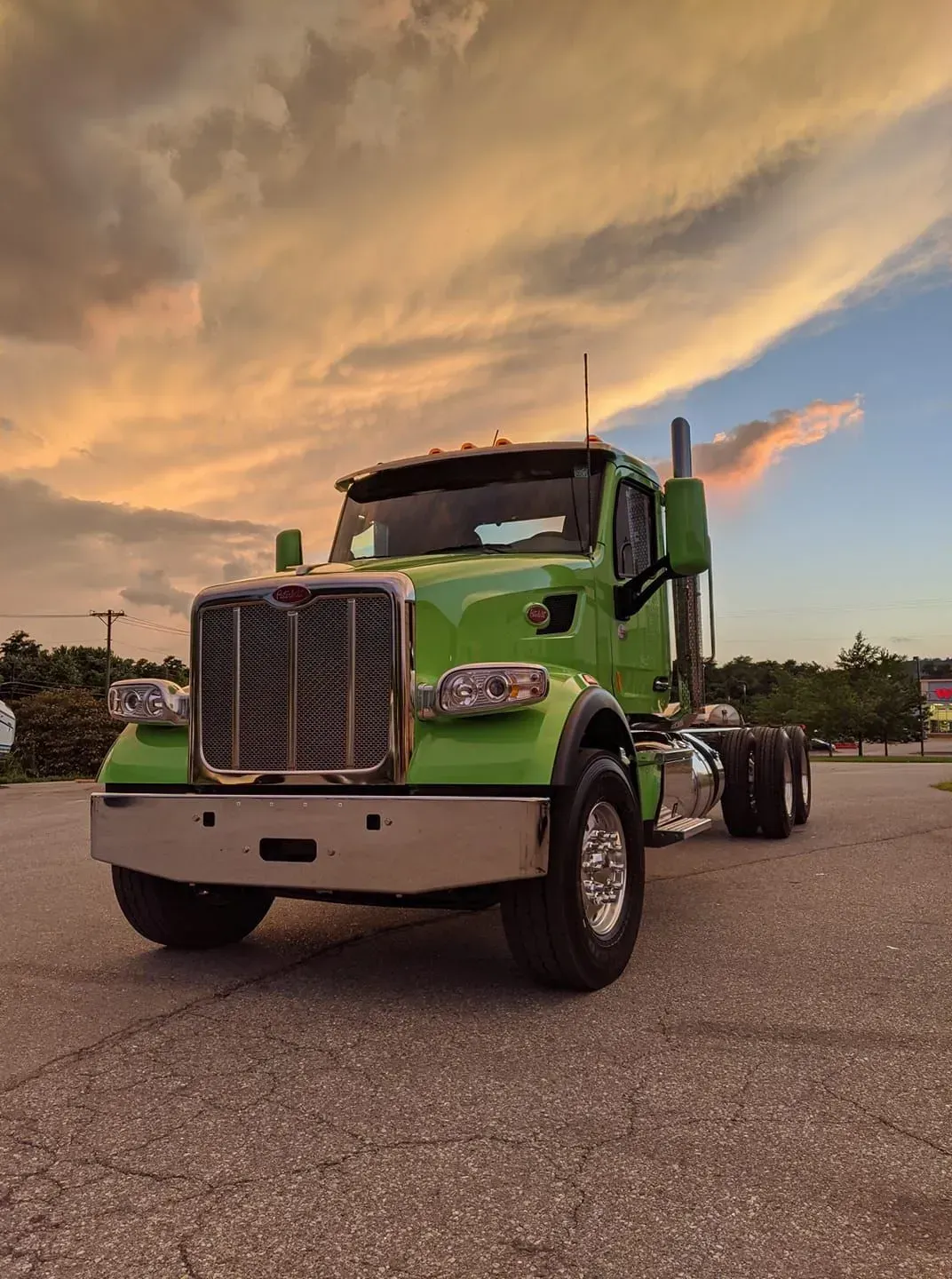 Green semi-truck parked on asphalt, chrome details, under a colorful sunset.