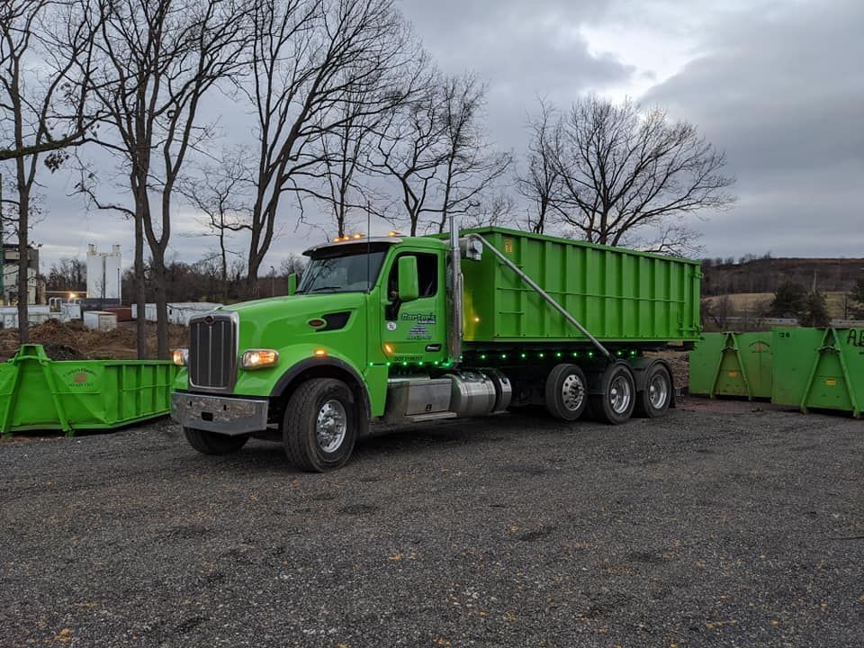 Green dump truck next to several green dumpsters, parked on gravel, overcast sky.
