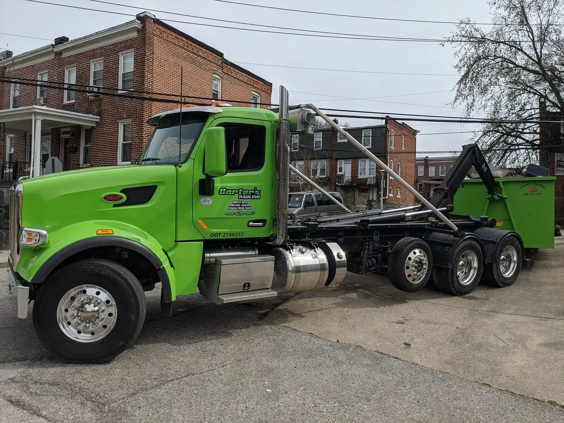 Green roll-off truck on a city street next to a brick building.