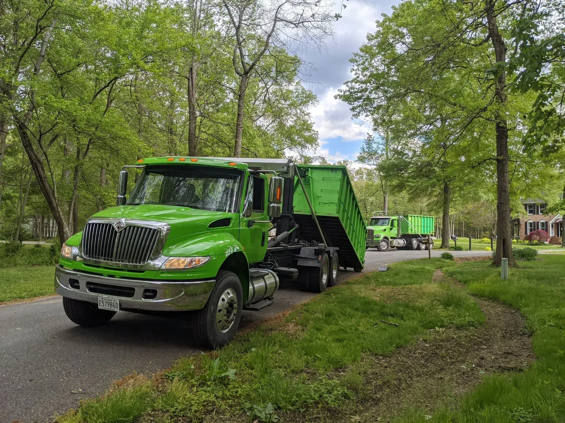 Green dump truck on a road, with a second truck visible in the distance, both in a residential area.