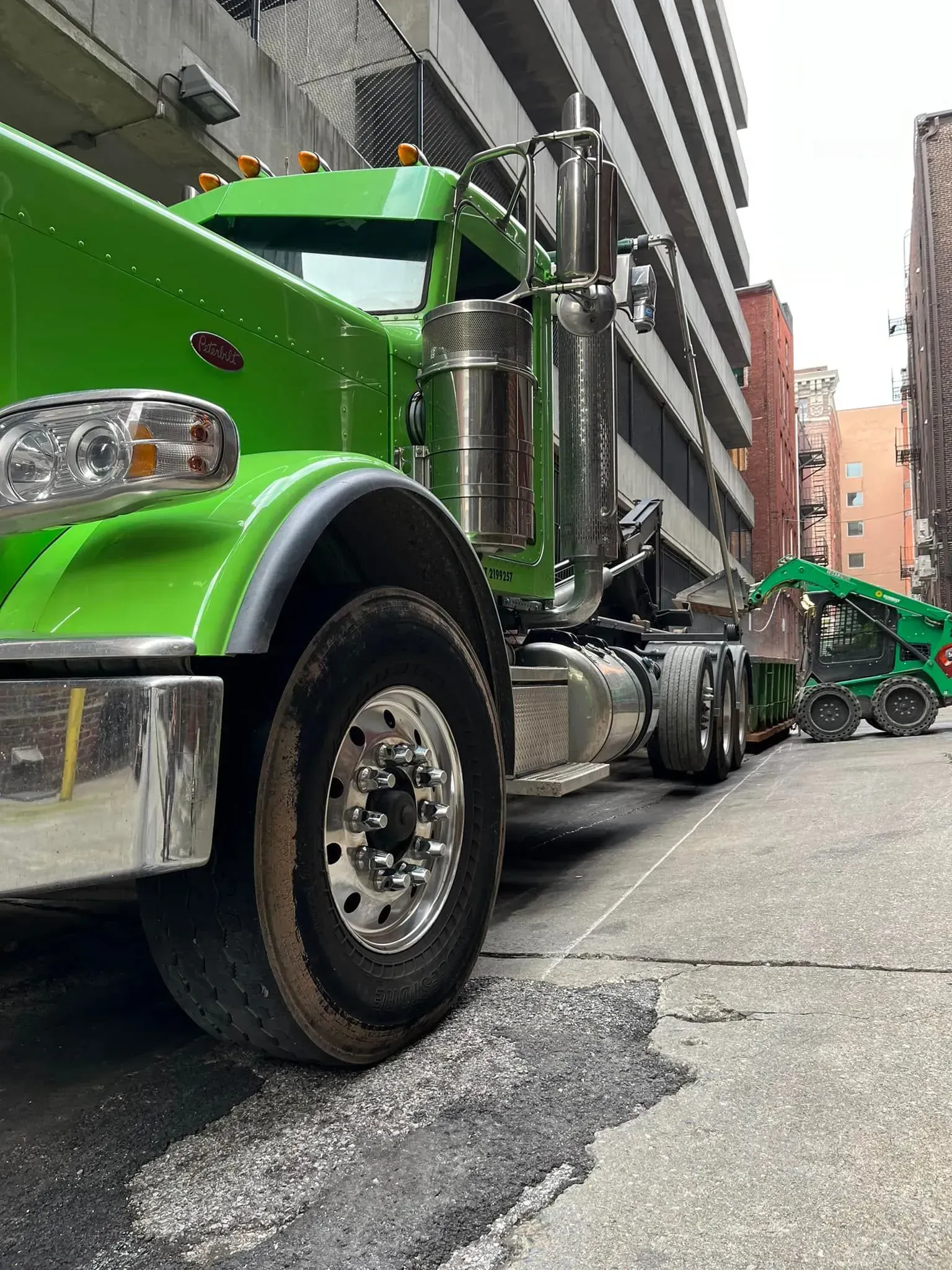 Green semi-truck in a narrow alley between buildings. Shiny chrome and a trailer in the distance.