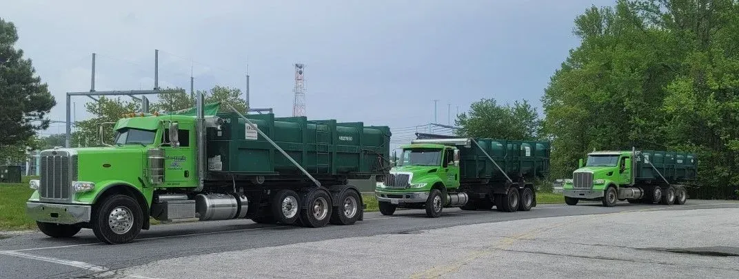 Three green dump trucks parked on a gravel lot next to trees.