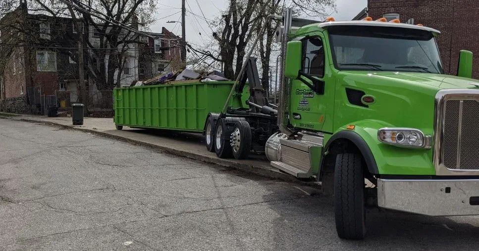 Green roll-off truck with container parked on a city street in front of a building.