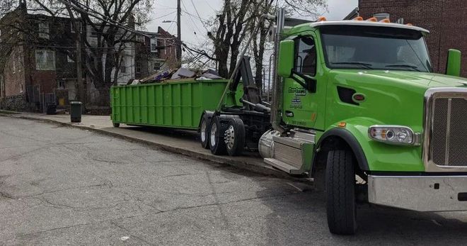 Green roll-off truck with container parked on a city street in front of a building.