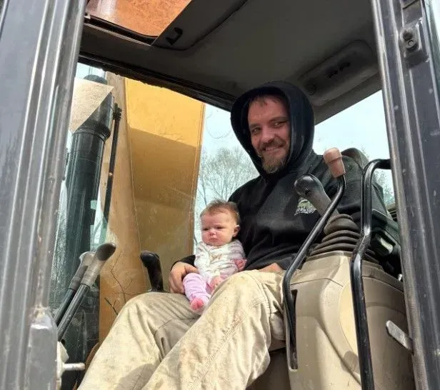 Man in hoodie holds baby in a heavy machinery cab. Both looking at the camera, smiles.