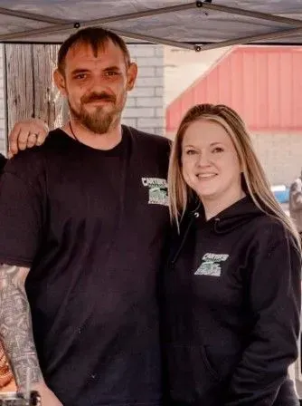 Man with arm tattoo and woman in matching black shirts, smiling outside.