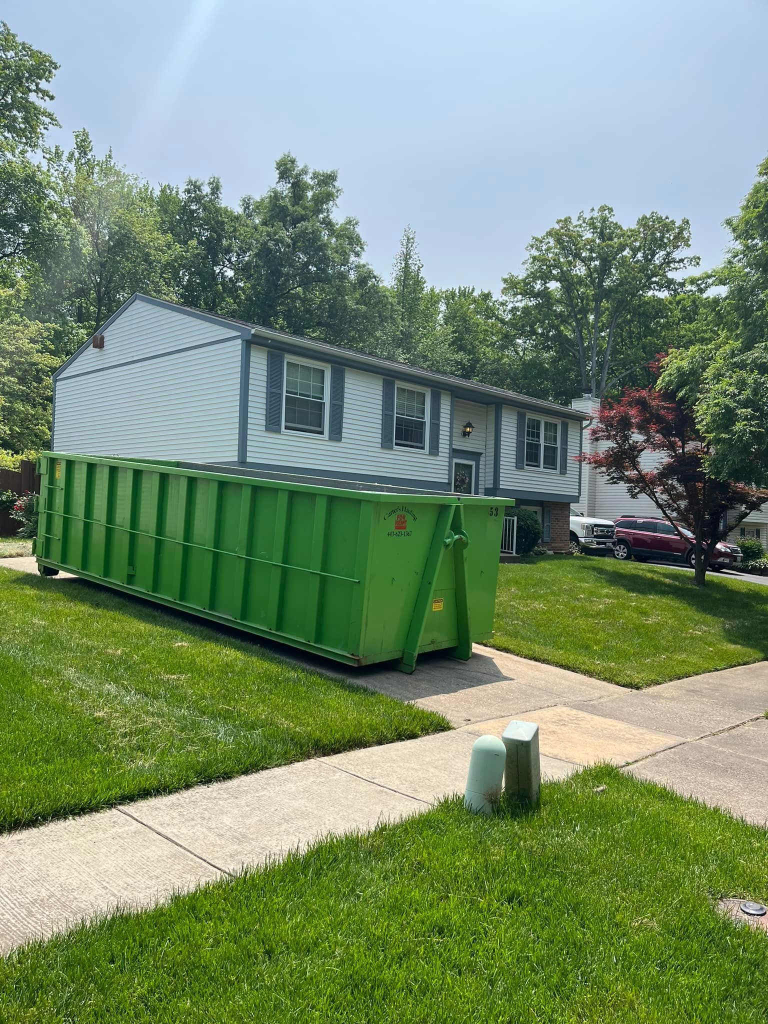 Green dumpster in front of a two-story house with lawn. Bright, sunny day.