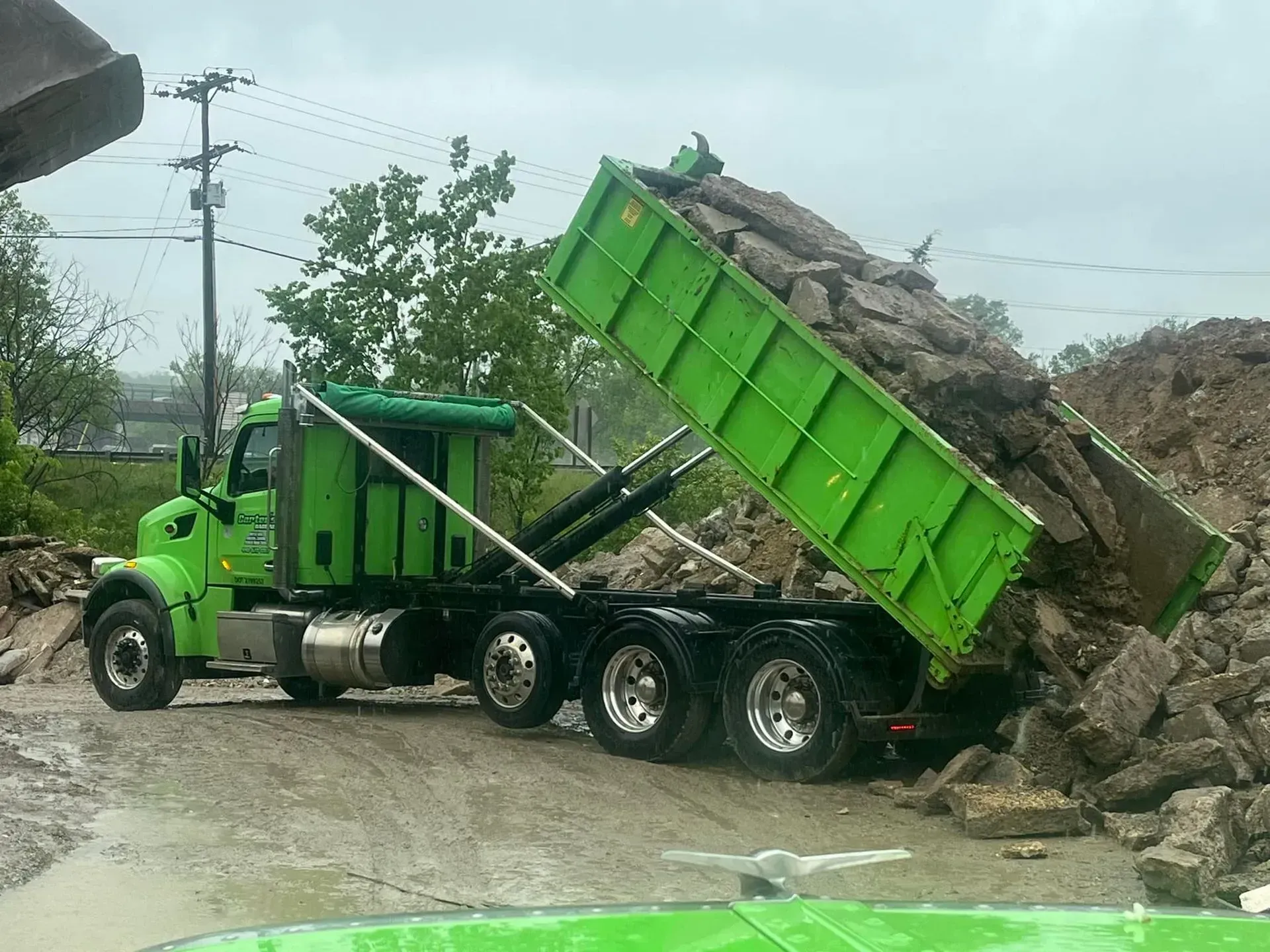 Green dump truck unloading rubble at a construction site, overcast day.