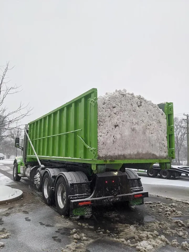 Green truck hauling a full load of snow on a snowy street.