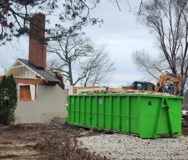 Demolished house with a green dumpster, excavator, and brick chimney. Cloudy sky.