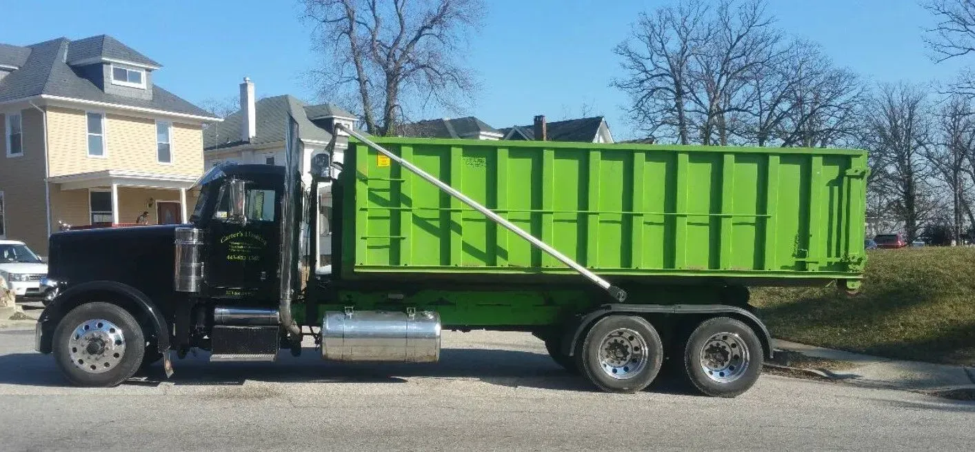 Black semi-truck with a bright green dumpster in front of a house on a sunny day.