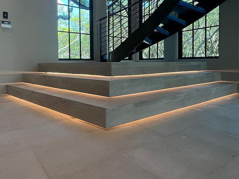 Stone steps with integrated warm lighting, leading to a staircase, in a building with large windows.