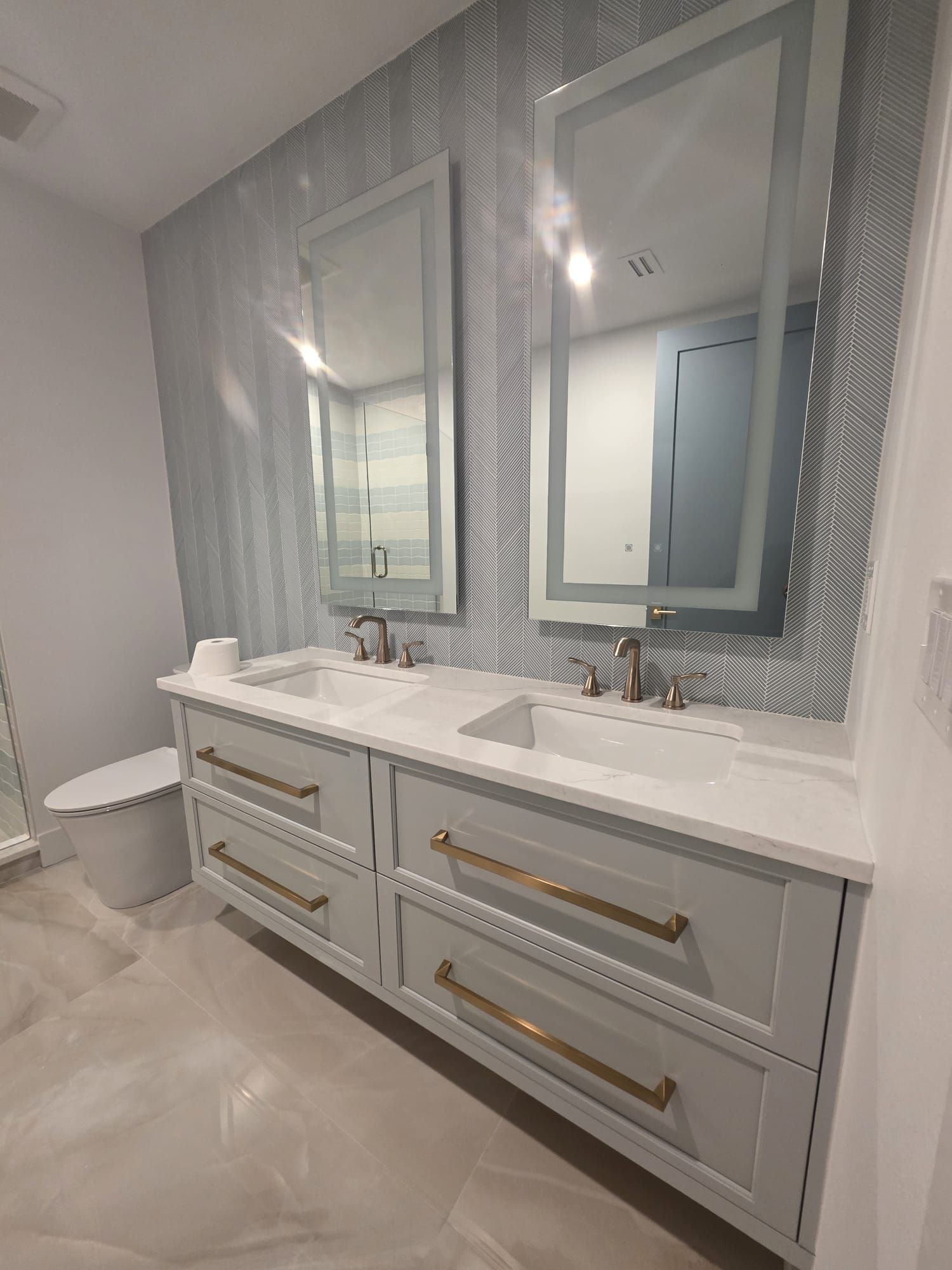 Double-sink bathroom vanity with mirrors, gray cabinets, gold handles, and patterned wallpaper.