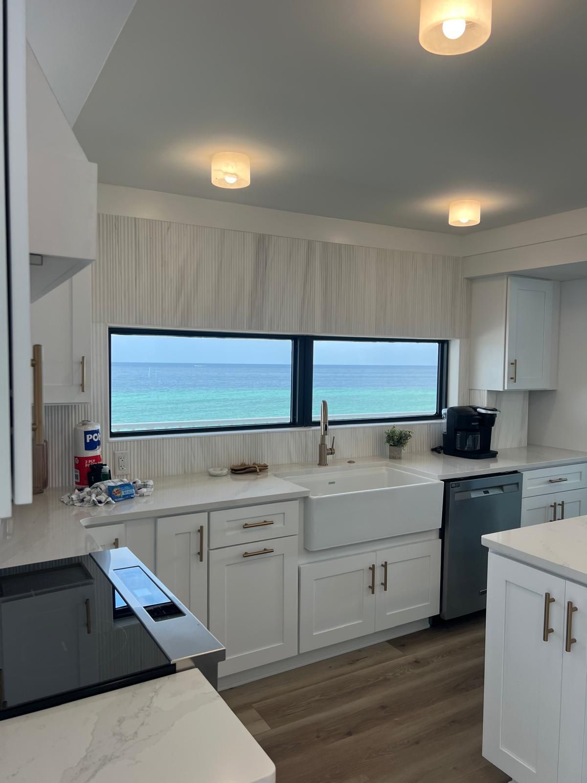 White kitchen with ocean view through a window, farmhouse sink, and light wooden floors.