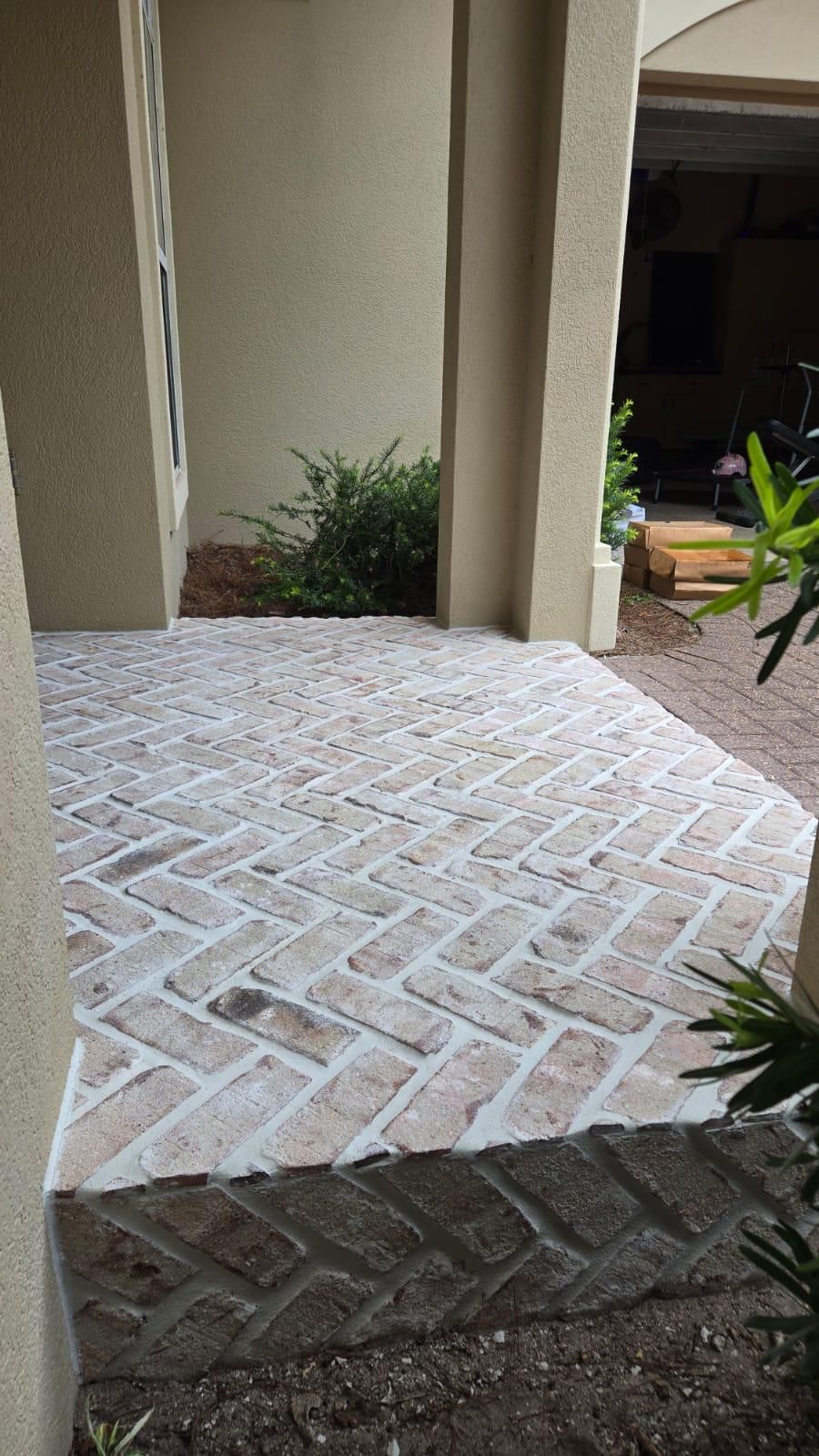 Herringbone brick entryway with light-colored bricks, beige stucco walls, and small green bush in the background.
