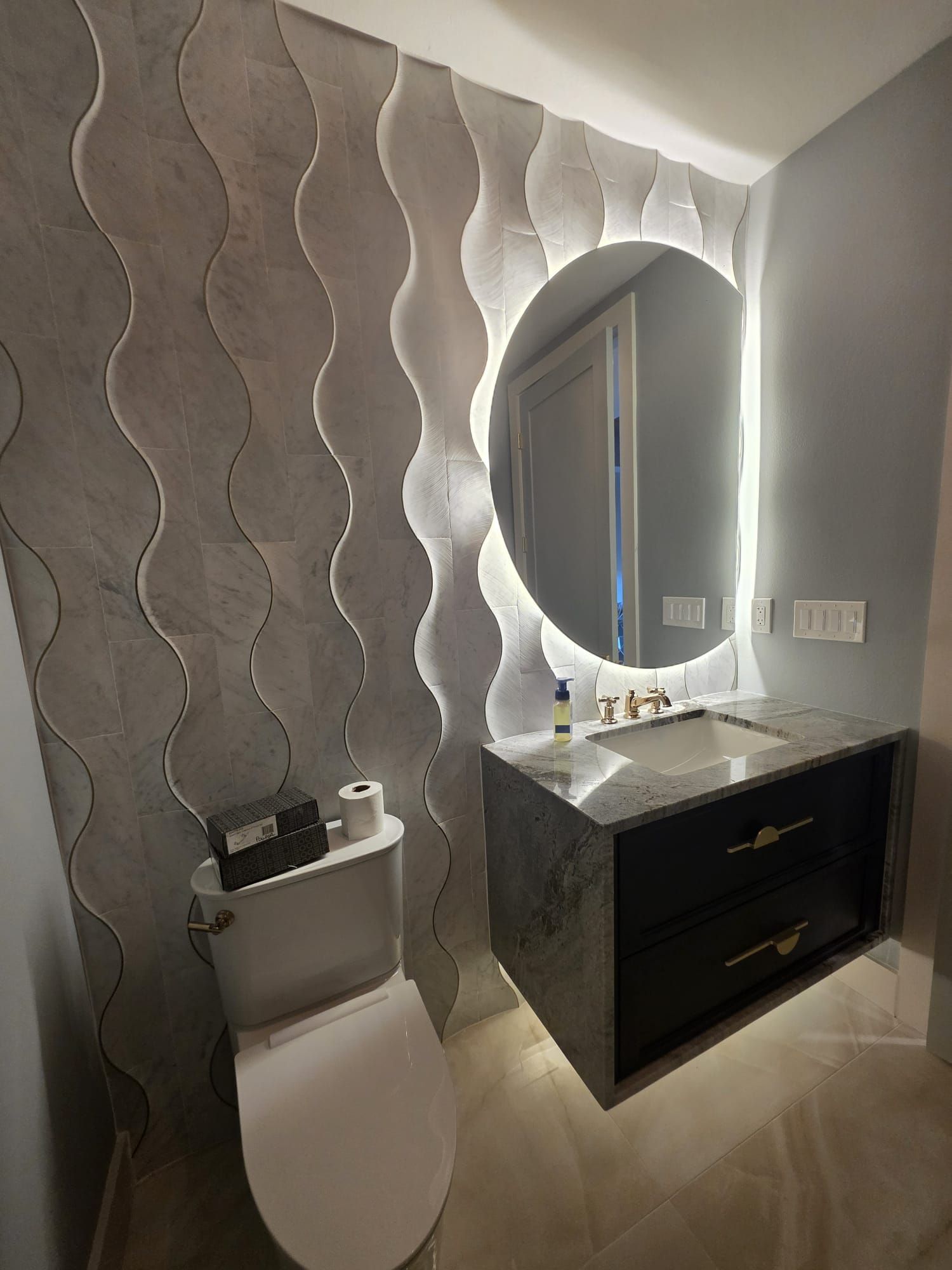 Modern bathroom with wavy gray wall tiles, floating vanity, and round illuminated mirror.