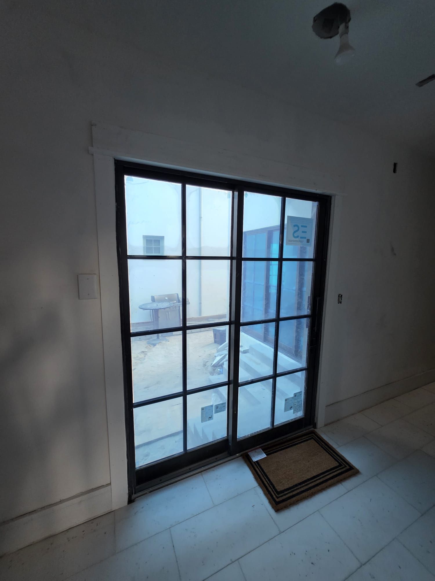 Black-framed glass door with multiple panes, revealing a construction site. A doormat sits in front of the door.