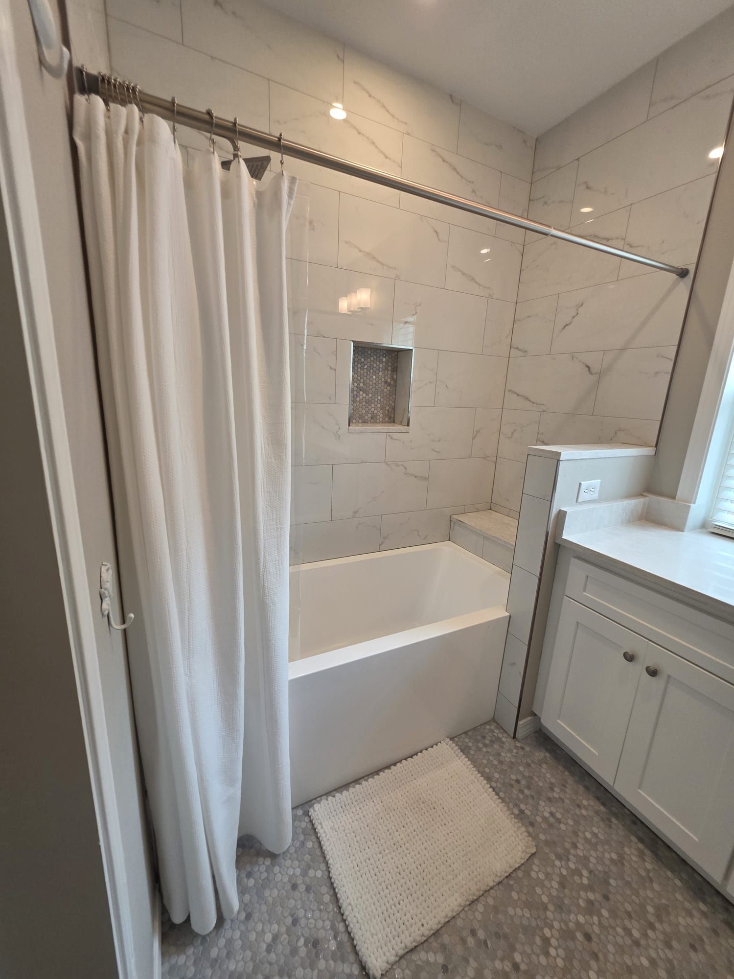 White-tiled bathroom with a bathtub, shower curtain, white cabinets, and a rug.