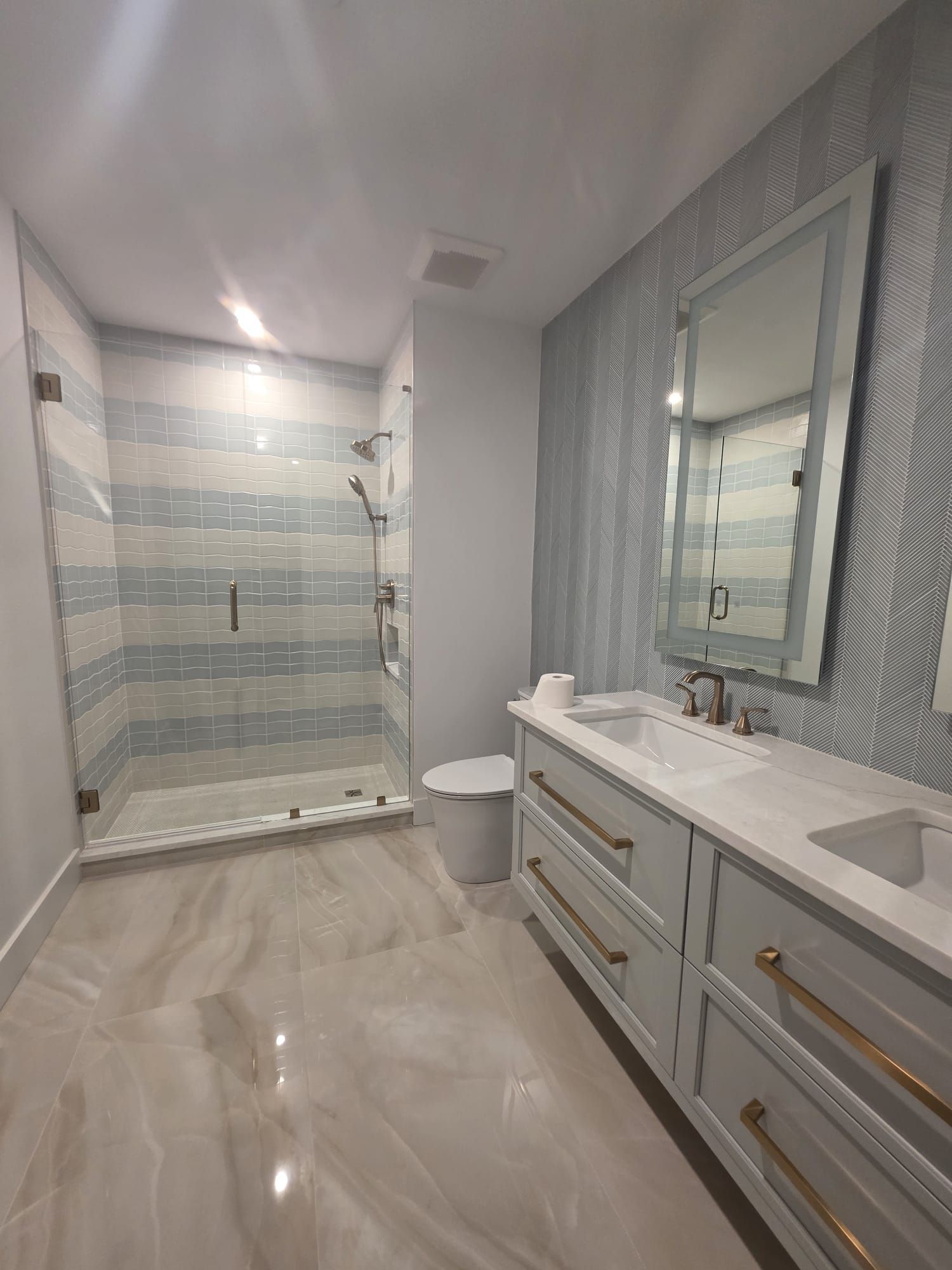 Modern bathroom with blue and white striped tile shower, double vanity, and light marble floor.