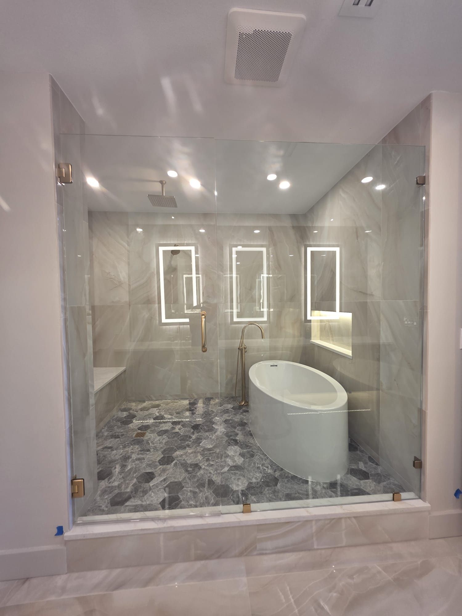 Glass-enclosed luxury bathroom with white soaking tub, marble flooring, and three illuminated mirrors.