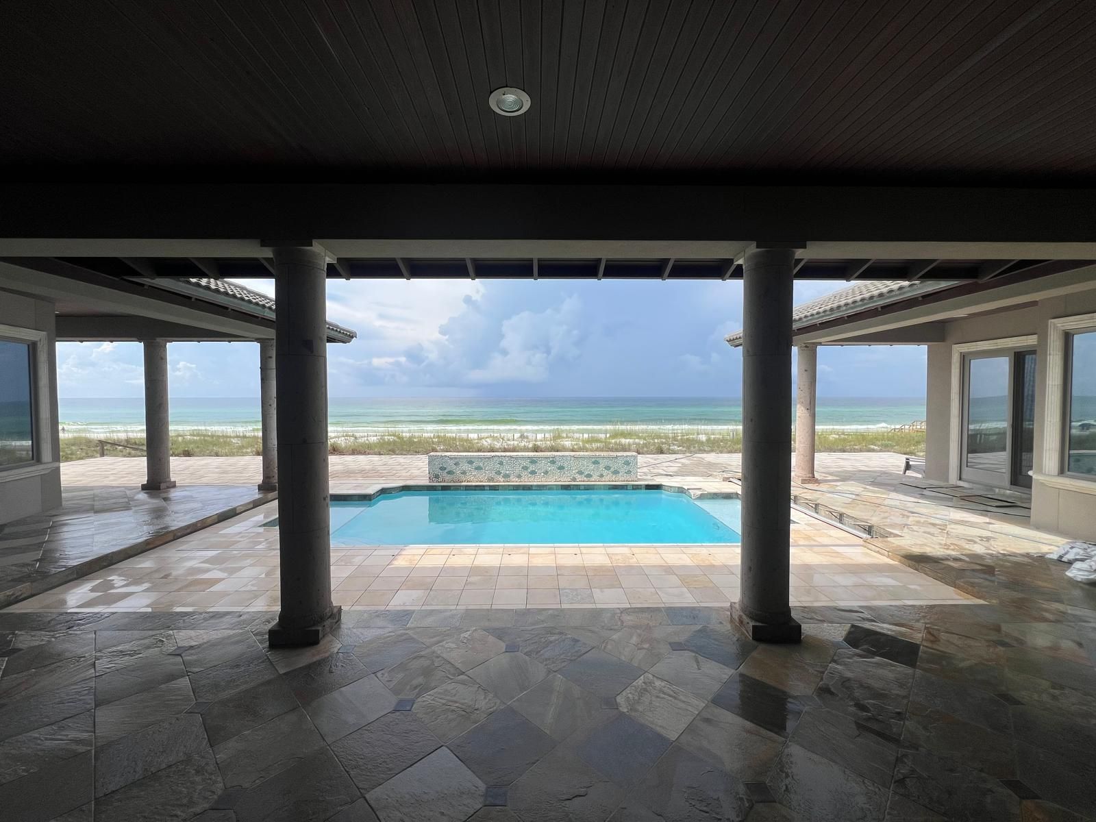 View of ocean and pool from a covered patio with pillars. Blue water and sandy beach.