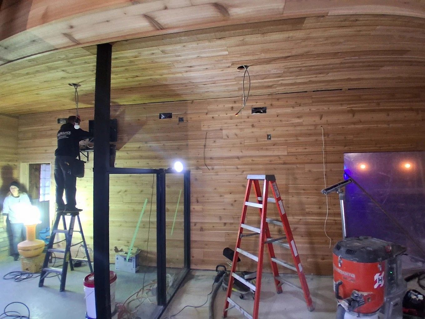 Workers installing a wood ceiling in a room with ladders, lights, and tools
