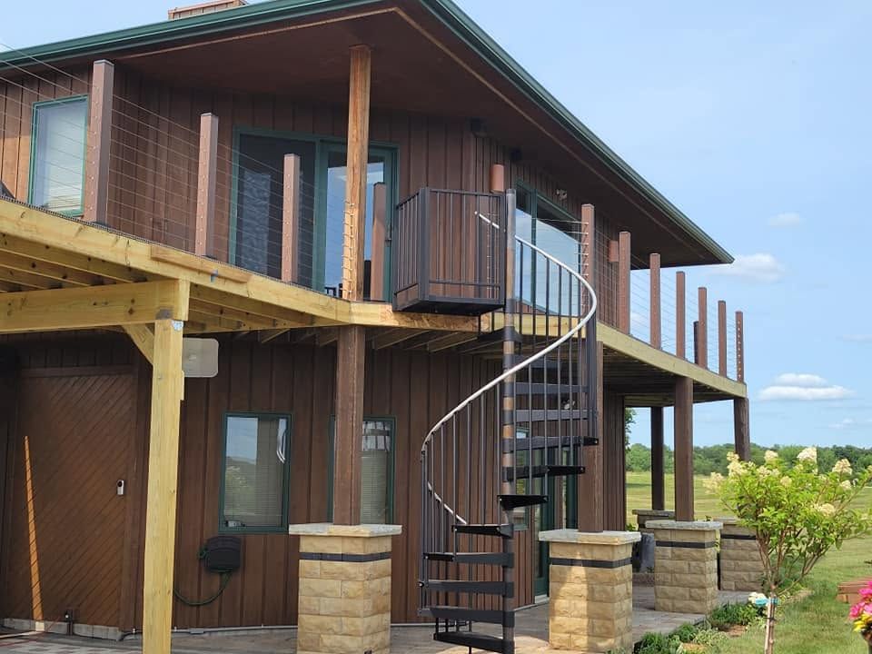 Two-story wooden house with a wraparound balcony and an outdoor spiral staircase under a blue sky