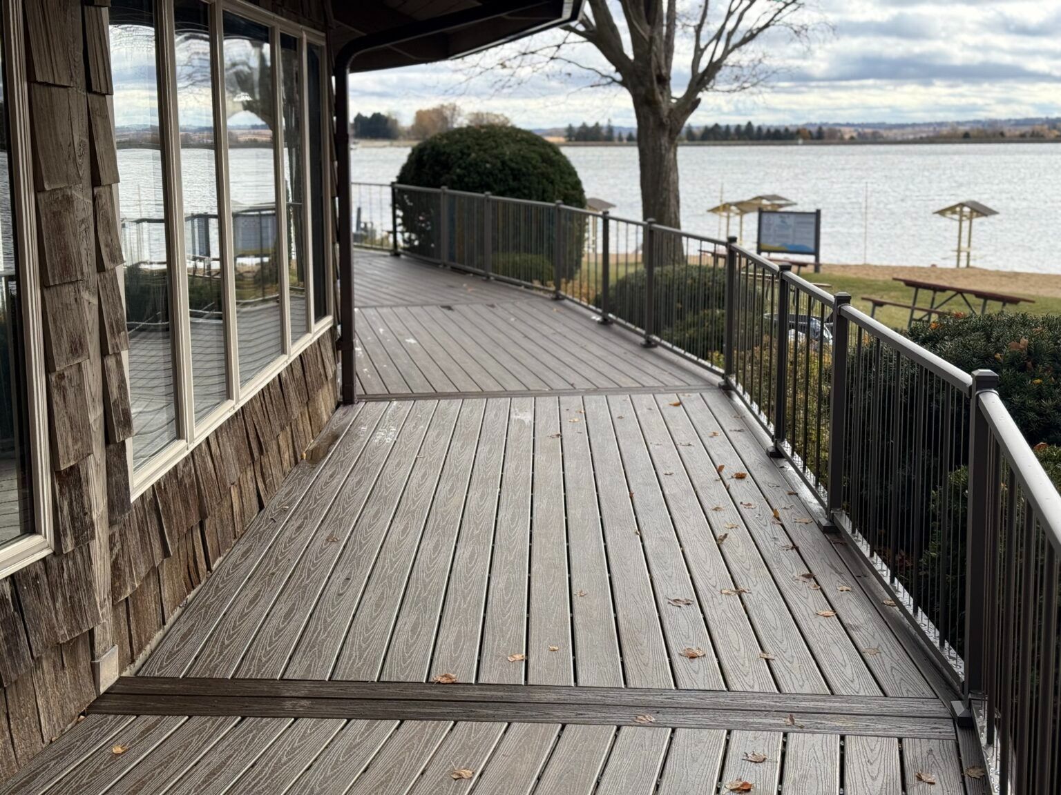Wooden deck walkway beside a waterfront building, with railing and trees overlooking the water.