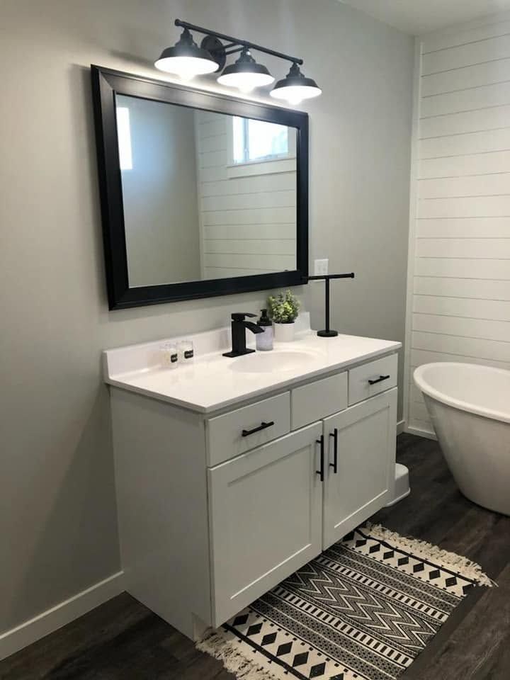 Modern bathroom vanity with black-framed mirror, double lights, white cabinets, and a patterned rug.
